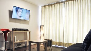 Un salón con televisión montada en la pared, una mesa de madera, una silla roja, un sofá negro y una ventana con cortinas.