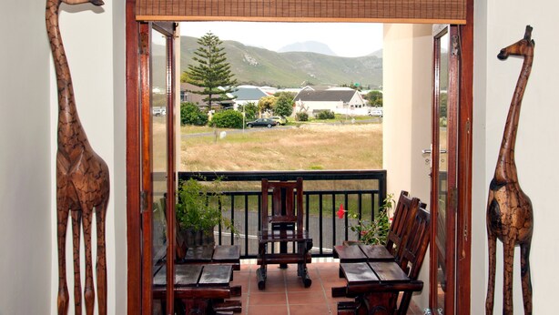 Ein Balkon mit Holzmöbeln und Giraffenskulpturen, der auf eine Wiese mit Häusern und Bergen blickt.