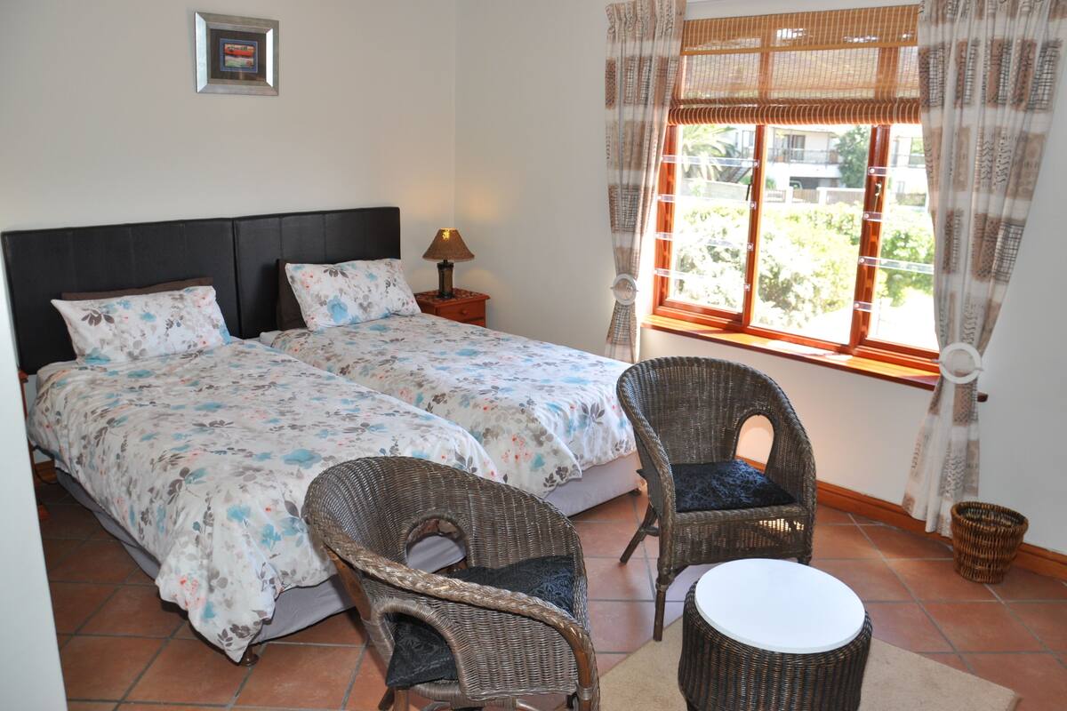 A double bed with floral bedding, two wicker chairs, a small table, and a window with curtains.