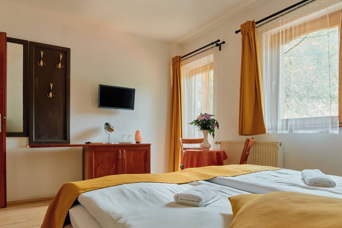 A hotel room with a bed, a TV, a wooden cabinet, a table with a vase, and a window with curtains.