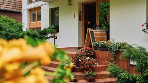 A building with a wooden door, a sign above the door, and a wooden barrel outside. There are steps leading up to the door, and potted plants are placed around the steps. - Oko-Park Panzió Kemping és Rendezvényközpont (Szarvasko)