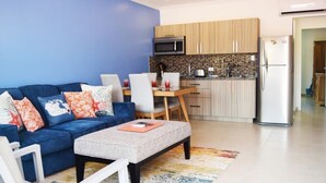 Uma sala de estar moderna com um sofá azul, uma mesa de centro e uma área de jantar com mesa e cadeiras de madeira. É visível, ao fundo, uma cozinha com armários de madeira e uma geladeira.