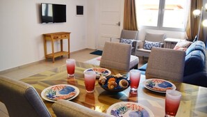Uma área de refeições com uma mesa posta para quatro pessoas, rodeada por cadeiras, uma televisão fixada na parede e um sofá com almofadas.