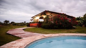 A swimming pool with a wooden deck leading to a house with a sloped roof and a wind turbine. - Oh Linda Pousada (Icapuí)