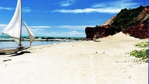 A sandy beach with a sailboat and rocky cliffs in the background. - Oh Linda Pousada (Icapuí)