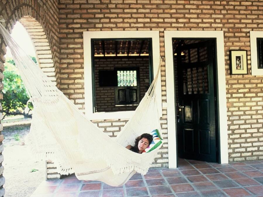 Una persona descansando en una hamaca sujeta a un edificio de ladrillo, con suelo de baldosas y un vano.