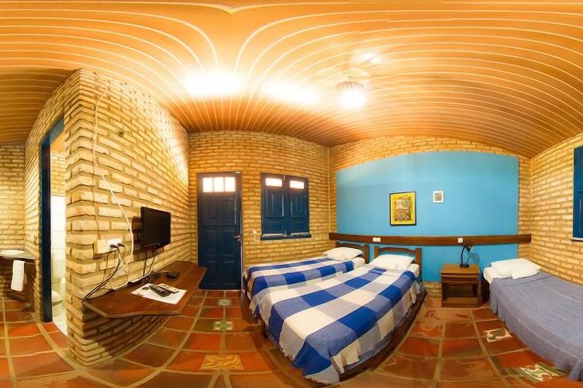 A room with brick walls, two beds with blue and white checkered bedding, a TV on a wooden stand, and a blue wall with a picture.