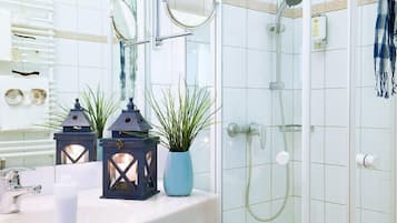 Une salle de bain avec un mur recouvert de carreaux blancs, un lavabo avec un miroir, deux lanternes, une plante en pot et une douche équipée d’un pommeau de douche à main.