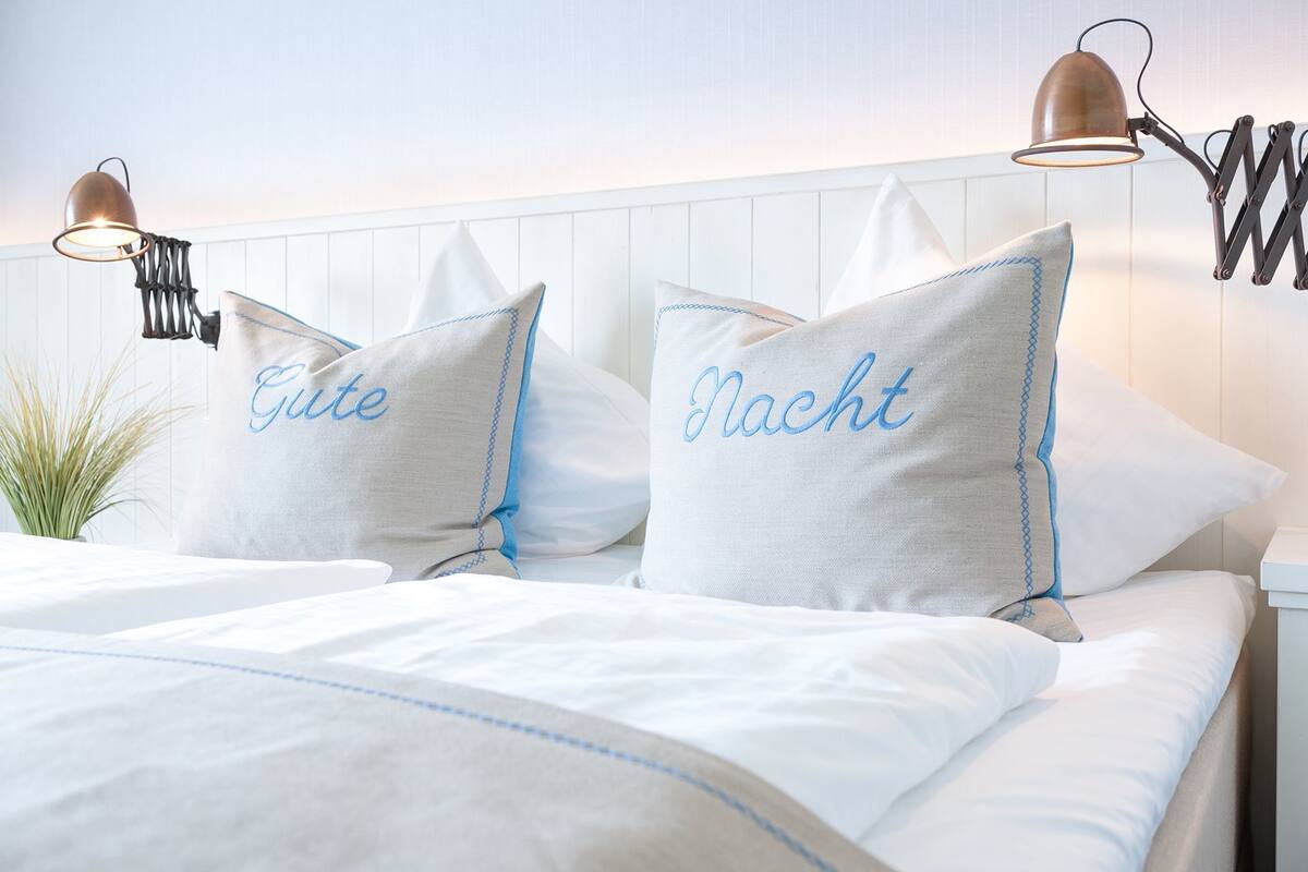 Een net opgemaakt bed met twee kussens, waarvan één het woord 'Gute' en de ander 'Nacht' heeft, plus twee wandlampen.