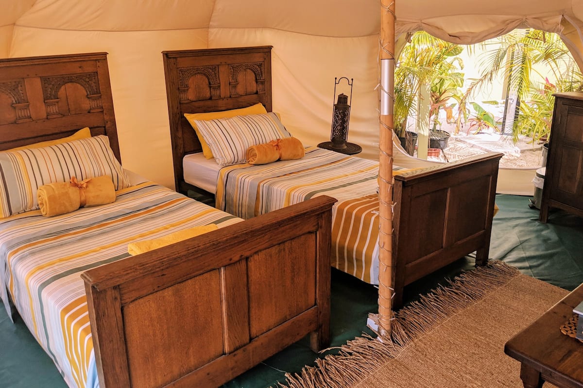 A tented accommodation with two wooden beds, each with striped bedding and pillows, a small table, and a hanging lantern.