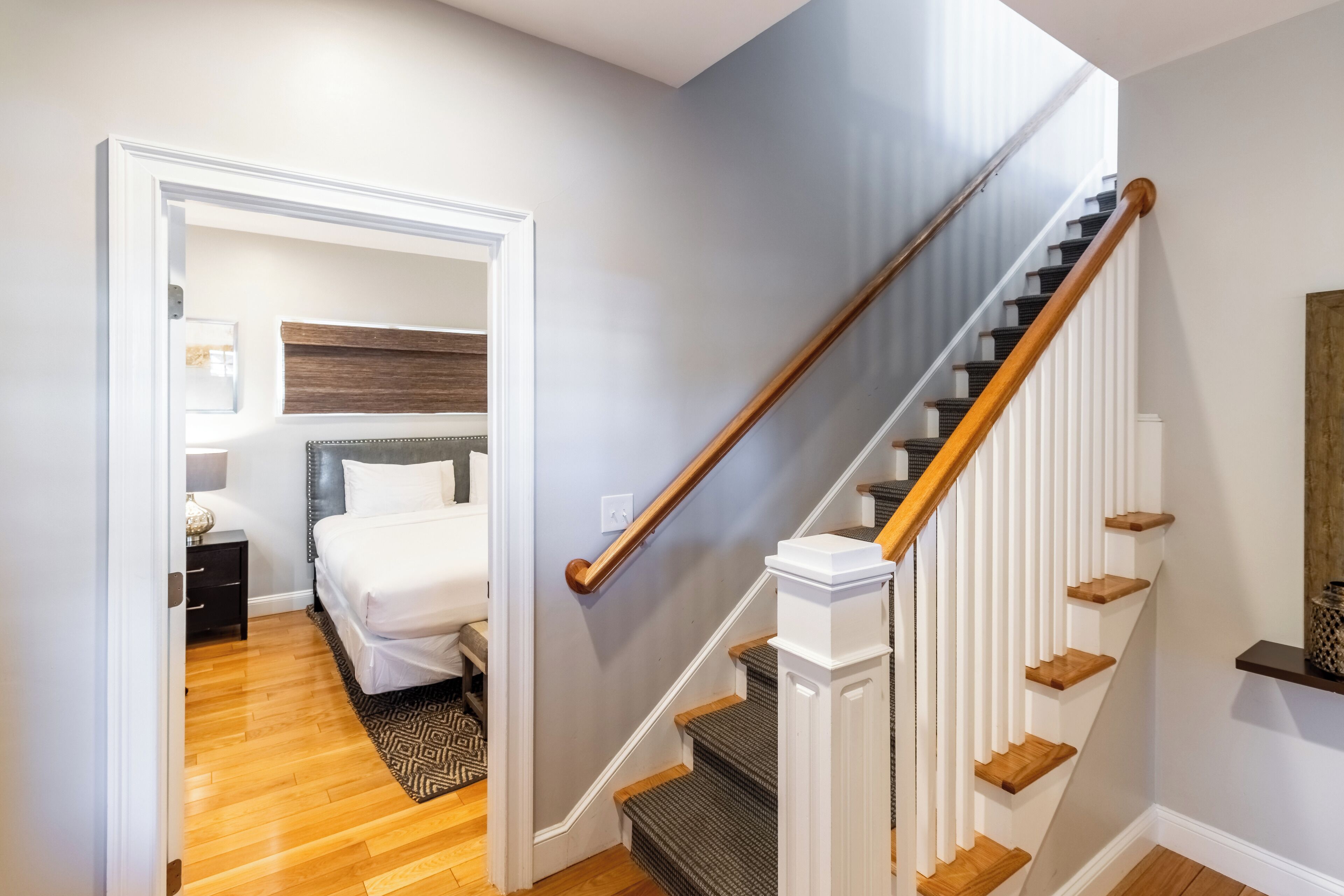 A bedroom with a bed, nightstand, and dresser, a staircase with wooden steps and white railings, and a hallway leading to the bedroom.