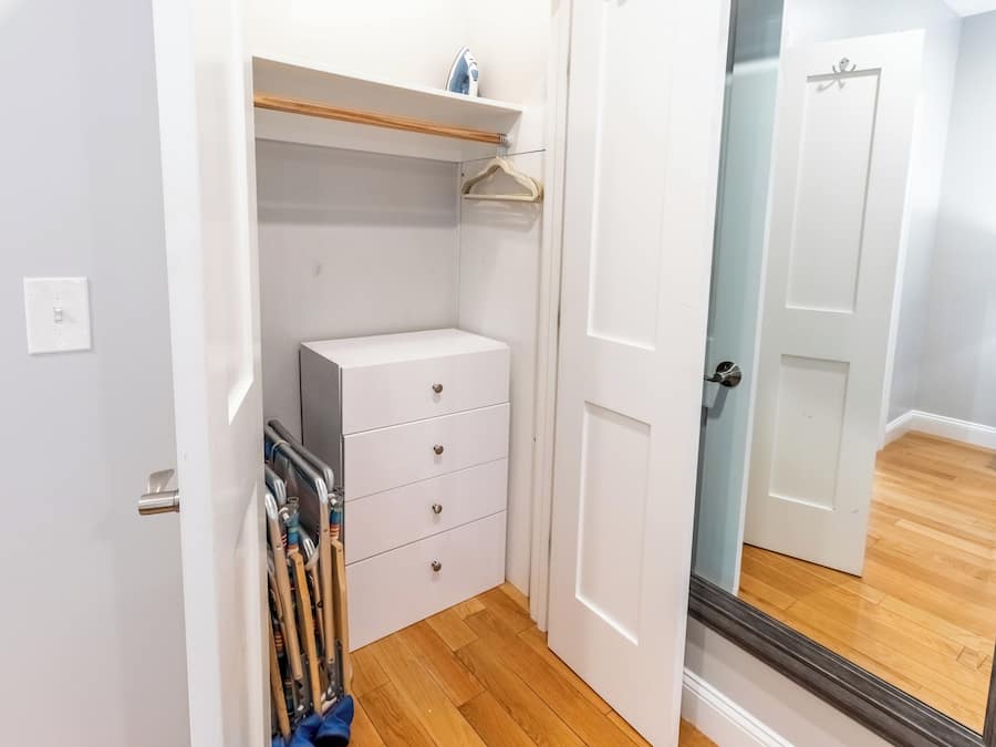 Un pasillo estrecho con una puerta blanca, piso de madera y un mueble blanco con cajones.