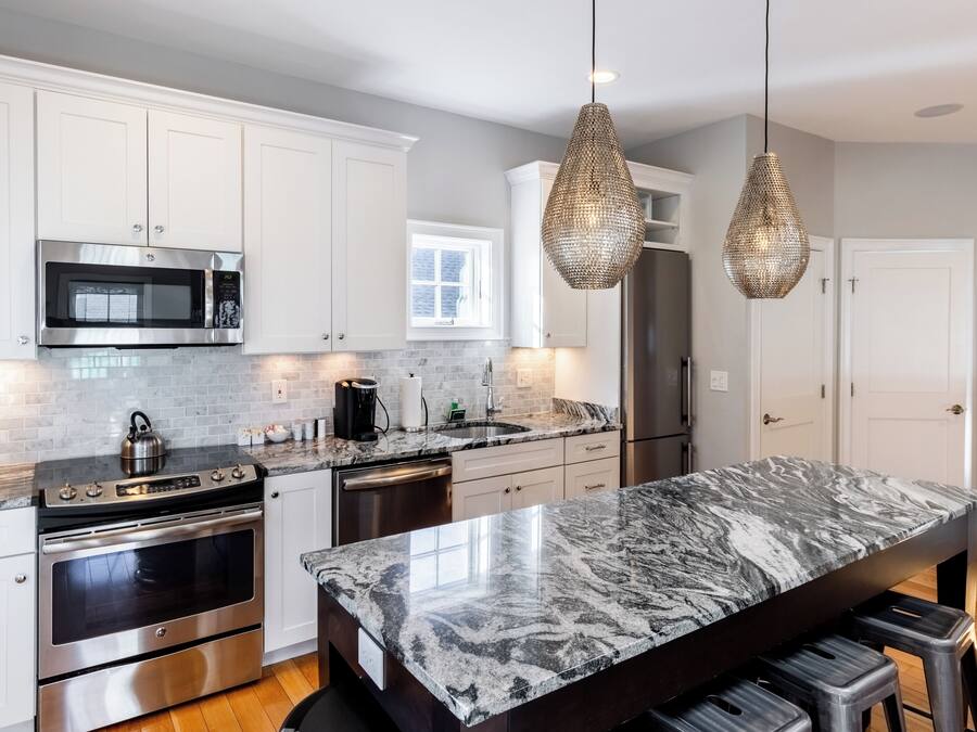 Una cocina moderna con armarios blancos, electrodomésticos de acero inoxidable, una isla de mármol e iluminación colgante.