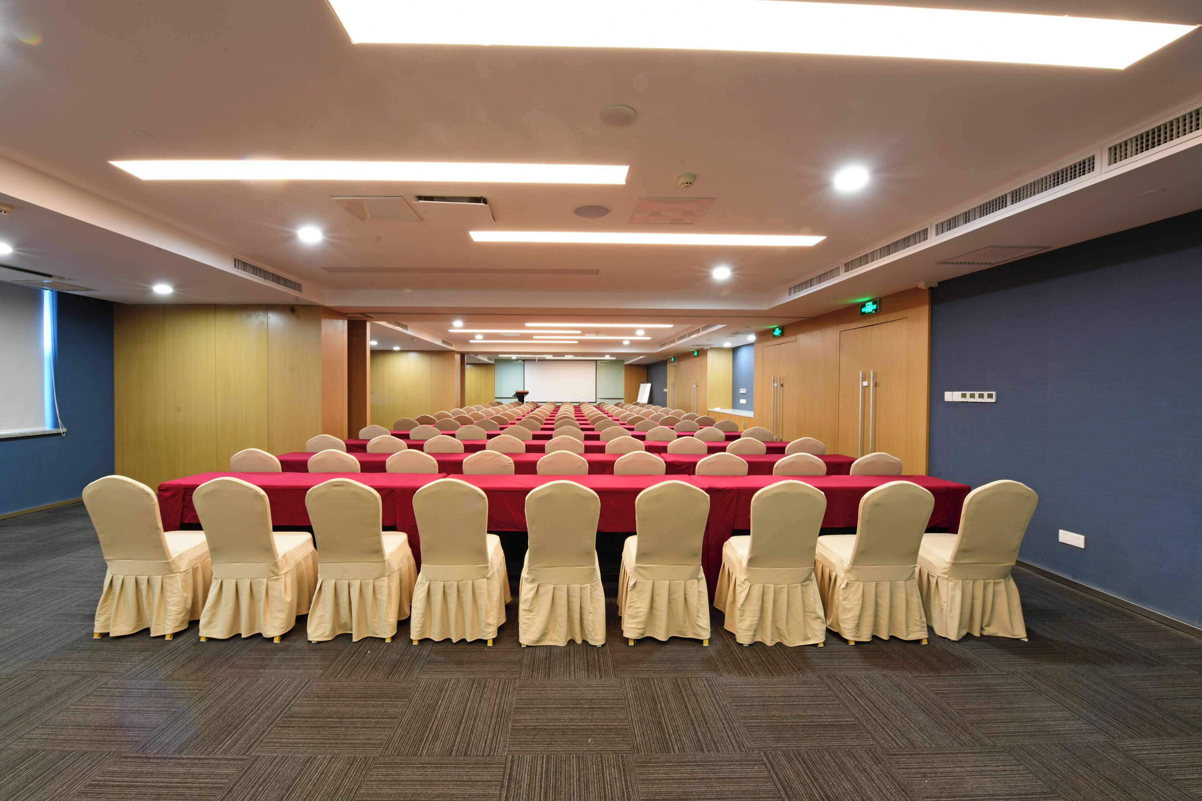 Un salón de conferencias con filas de mesas y sillas, equipado con luces en el techo y una pantalla de proyector.