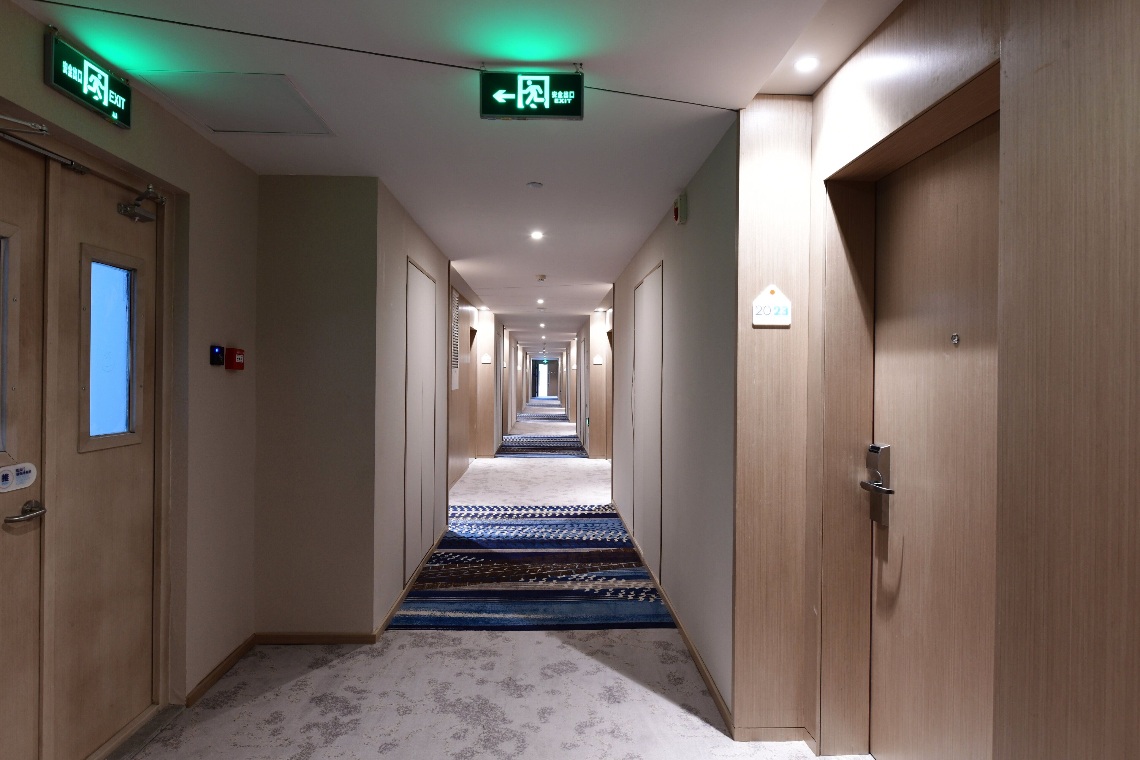 Un pasillo de hotel con pisos alfombrados, puertas de madera y letreros de salida.