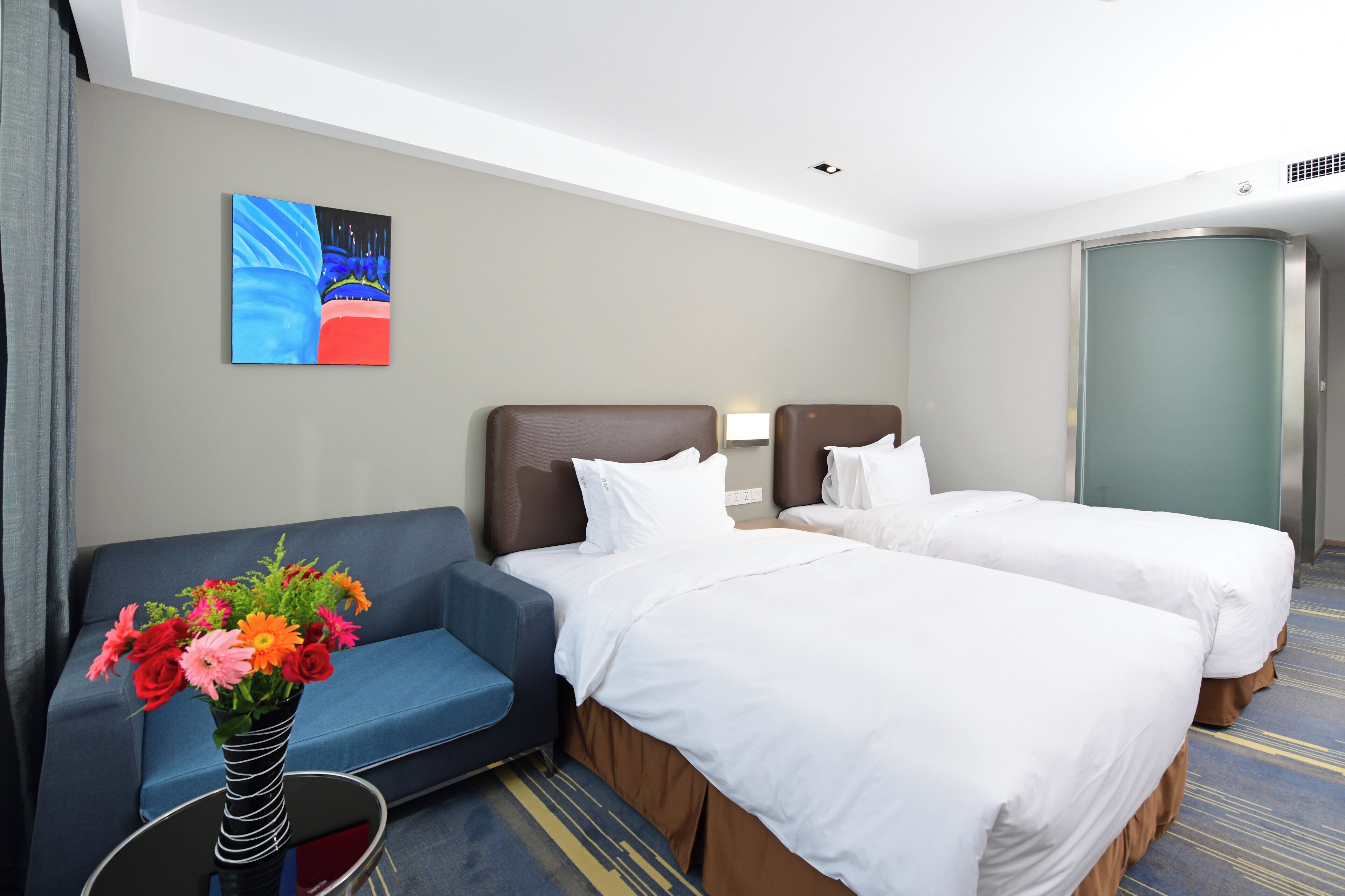 A hotel room with two beds, a sofa, a vase with flowers, and a painting on the wall.