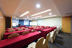 Un salón de conferencias con filas de mesas y sillas, una pizarra y una pantalla de presentación.