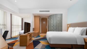 Quarto de hotel com cama, escrivaninha, cadeira, televisão e uma janela grande com cortinas.