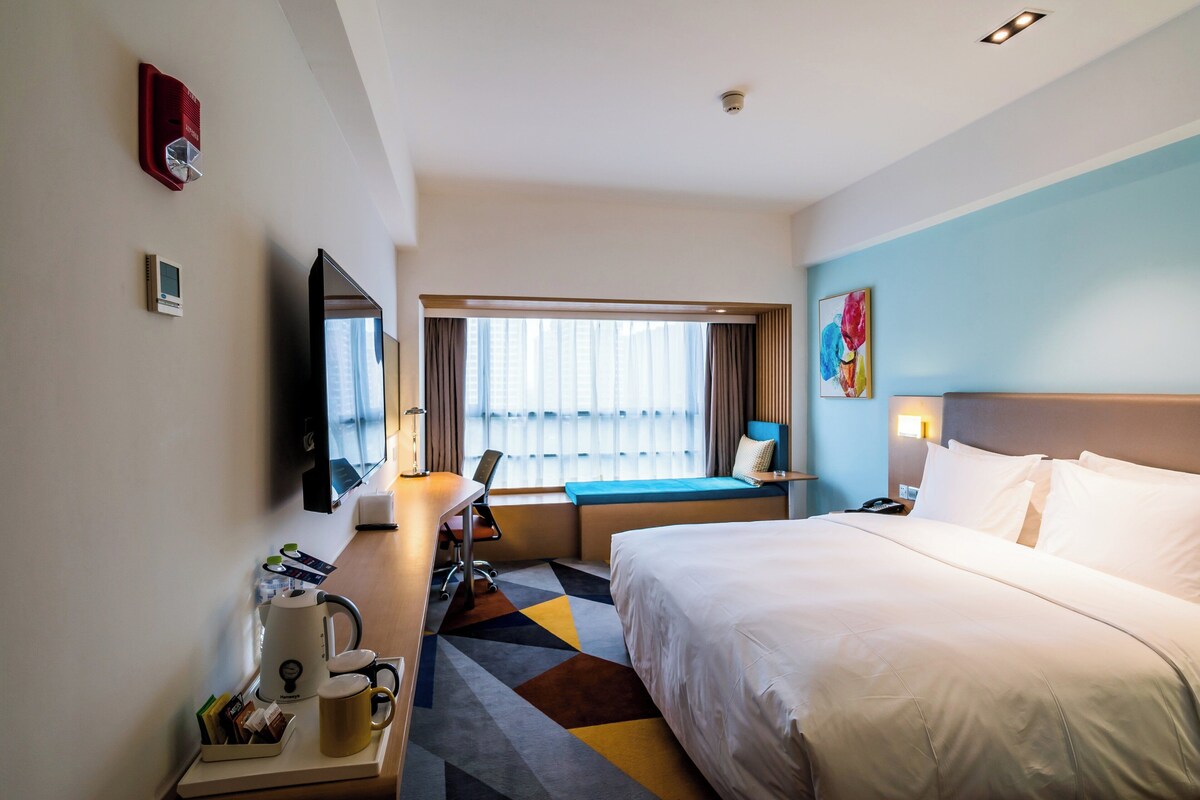 A hotel room with a bed, desk, chair, coffee maker, and a window with curtains.