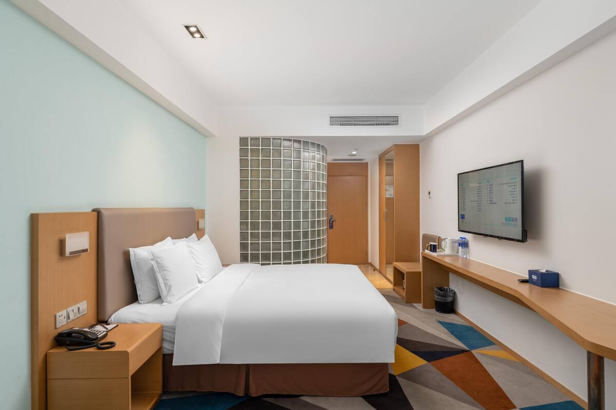 A hotel room with a bed, bedside table, a desk, a TV, and a wall-mounted screen.