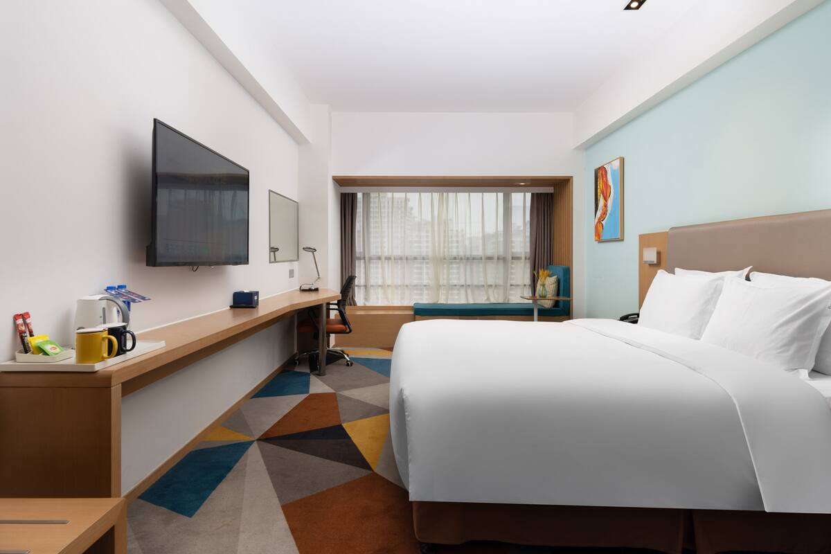 A hotel room with a bed, a desk, a flat-screen TV, a window with curtains, and a painting on the wall.