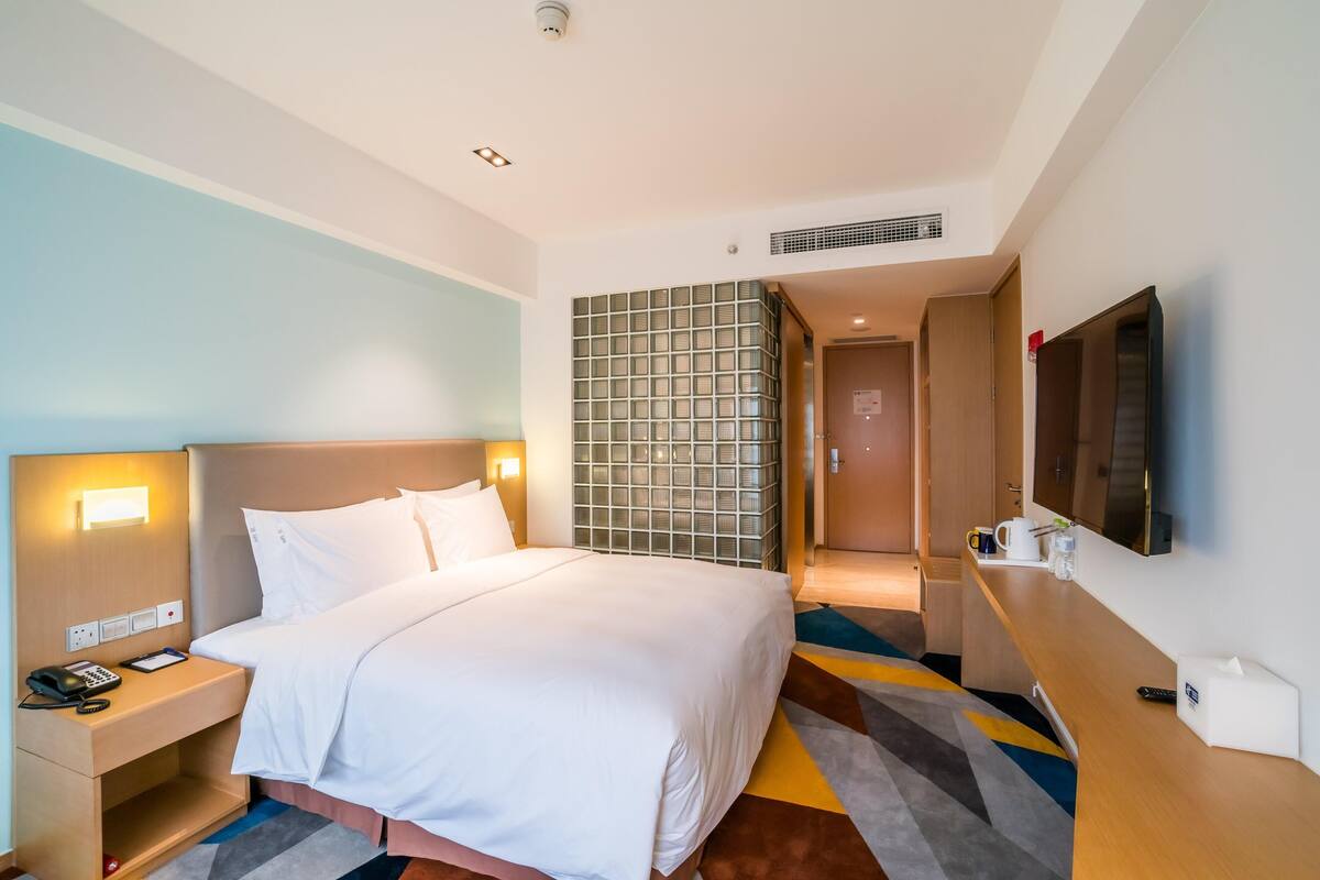 A hotel room with a large bed, bedside table with a phone and notepad, a TV mounted on the wall, and a patterned rug.