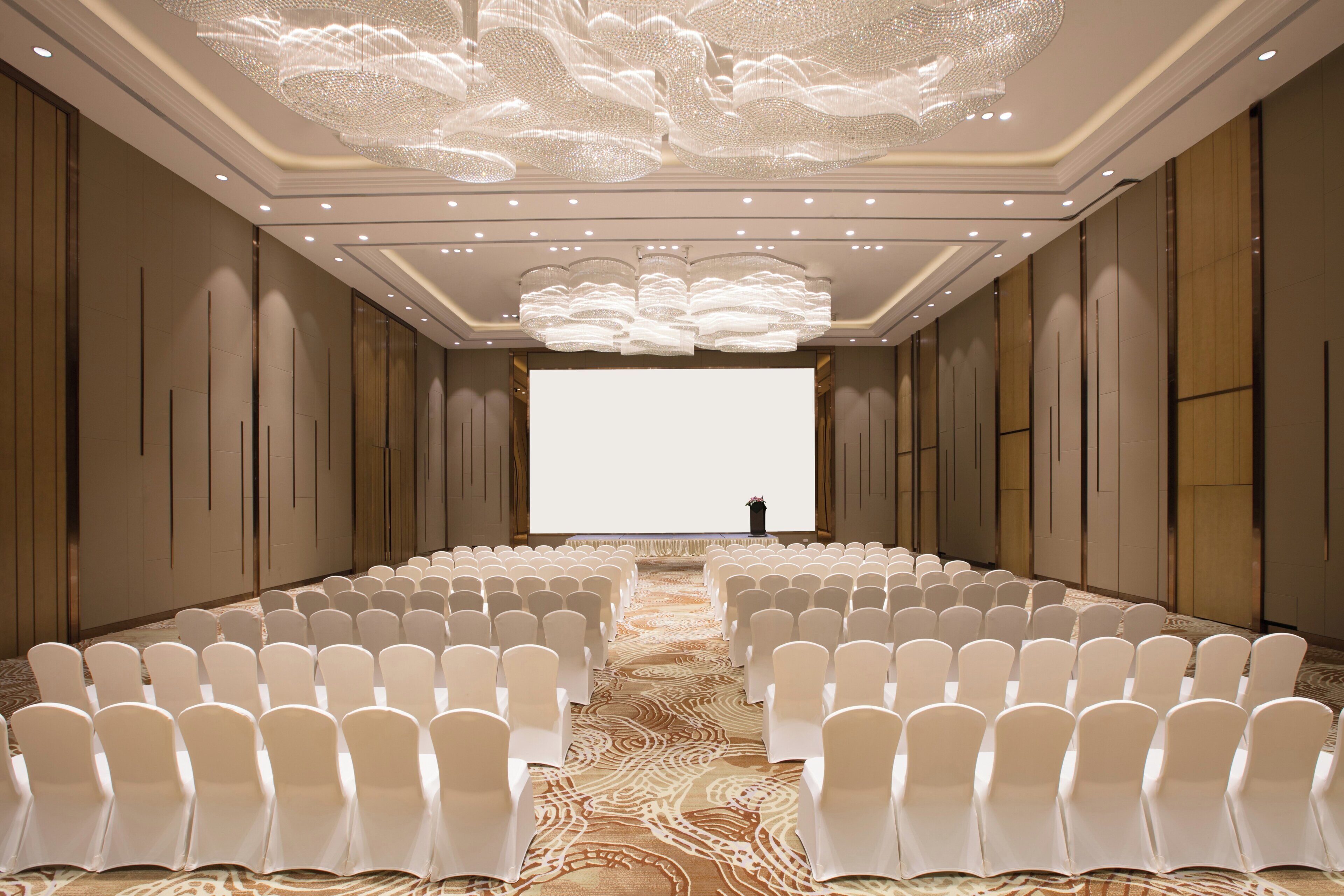 A conference room with rows of white chairs, a large screen at the front, and a decorative ceiling light fixture.