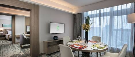 Una camera d'albergo moderna con zona pranzo, televisione e un'ampia finestra con tende.