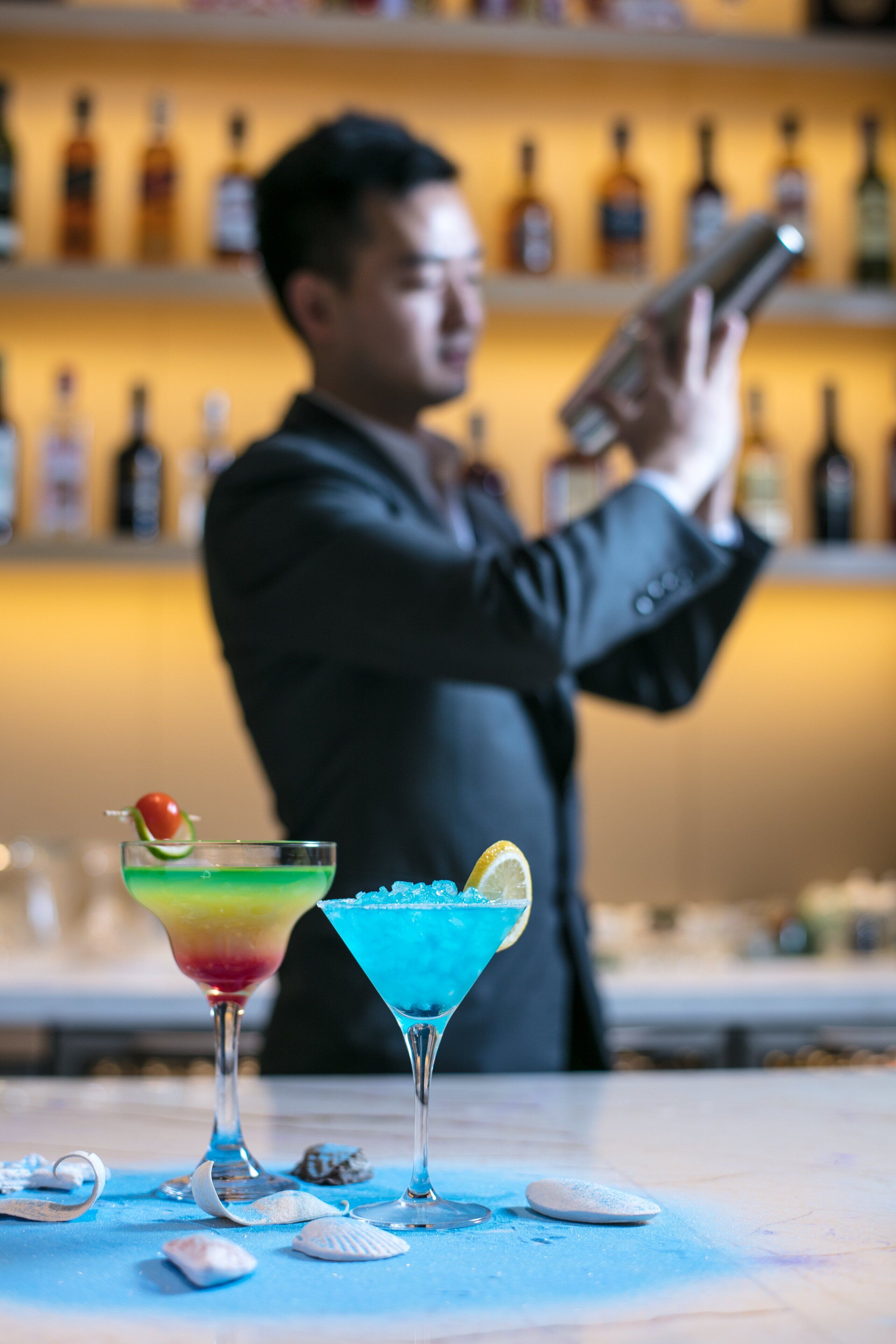A bartender preparing drinks at a bar with shelves of bottles in the background.
