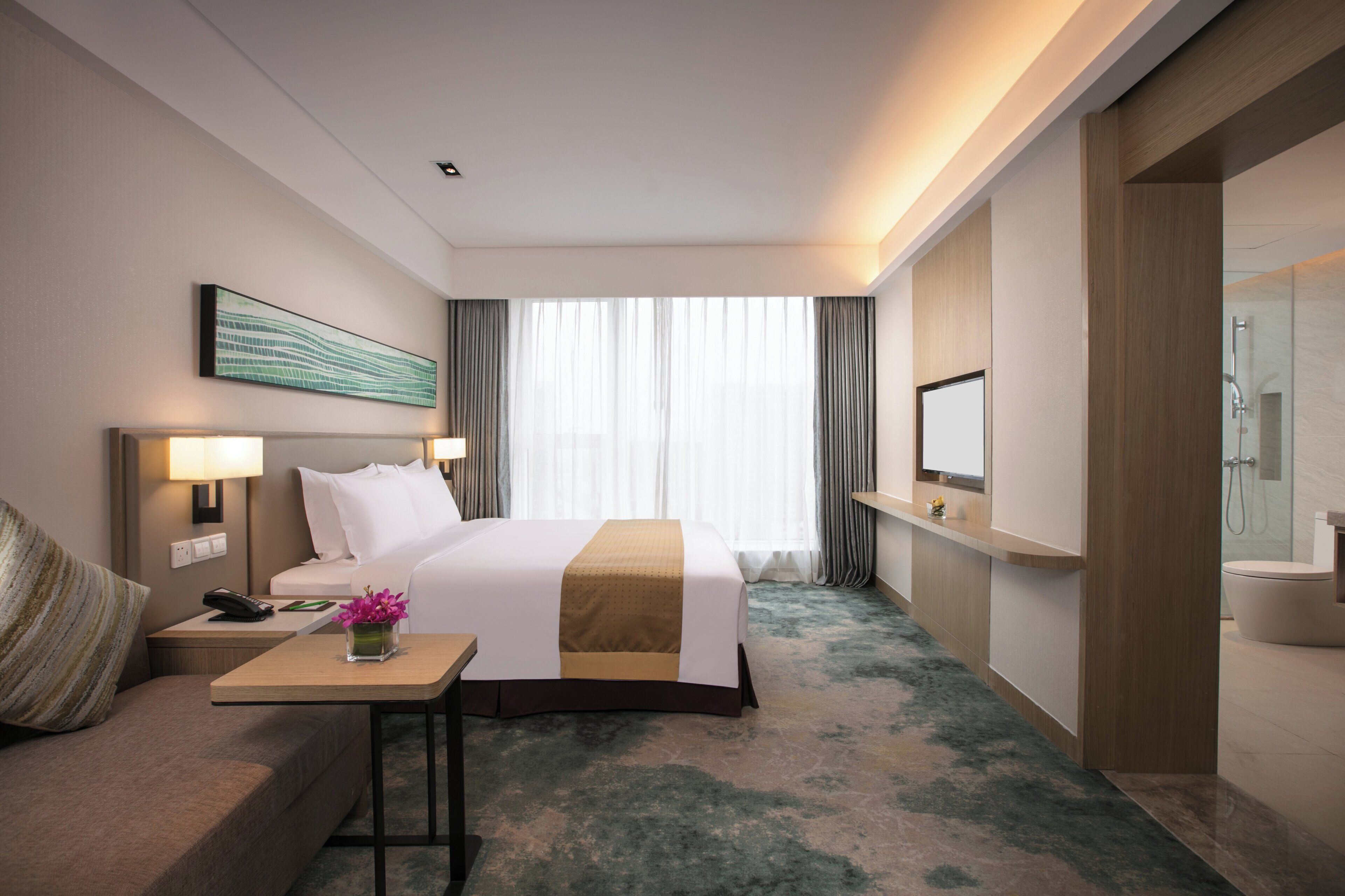 Habitación de hotel con cama, mesita de noche, lámpara y vistas al exterior por una ventana.