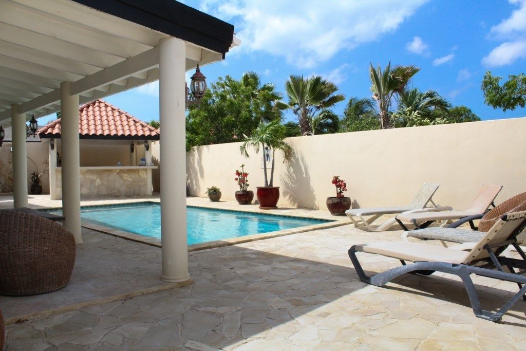 Zona de piscina con sillones reclinables, un porche cubierto y plantas en macetas.