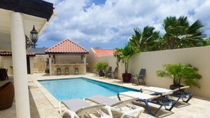 A pool area with lounge chairs, a tiled floor, and a building with a red-tiled roof in the background. - Casa Luna (Oranjestad)