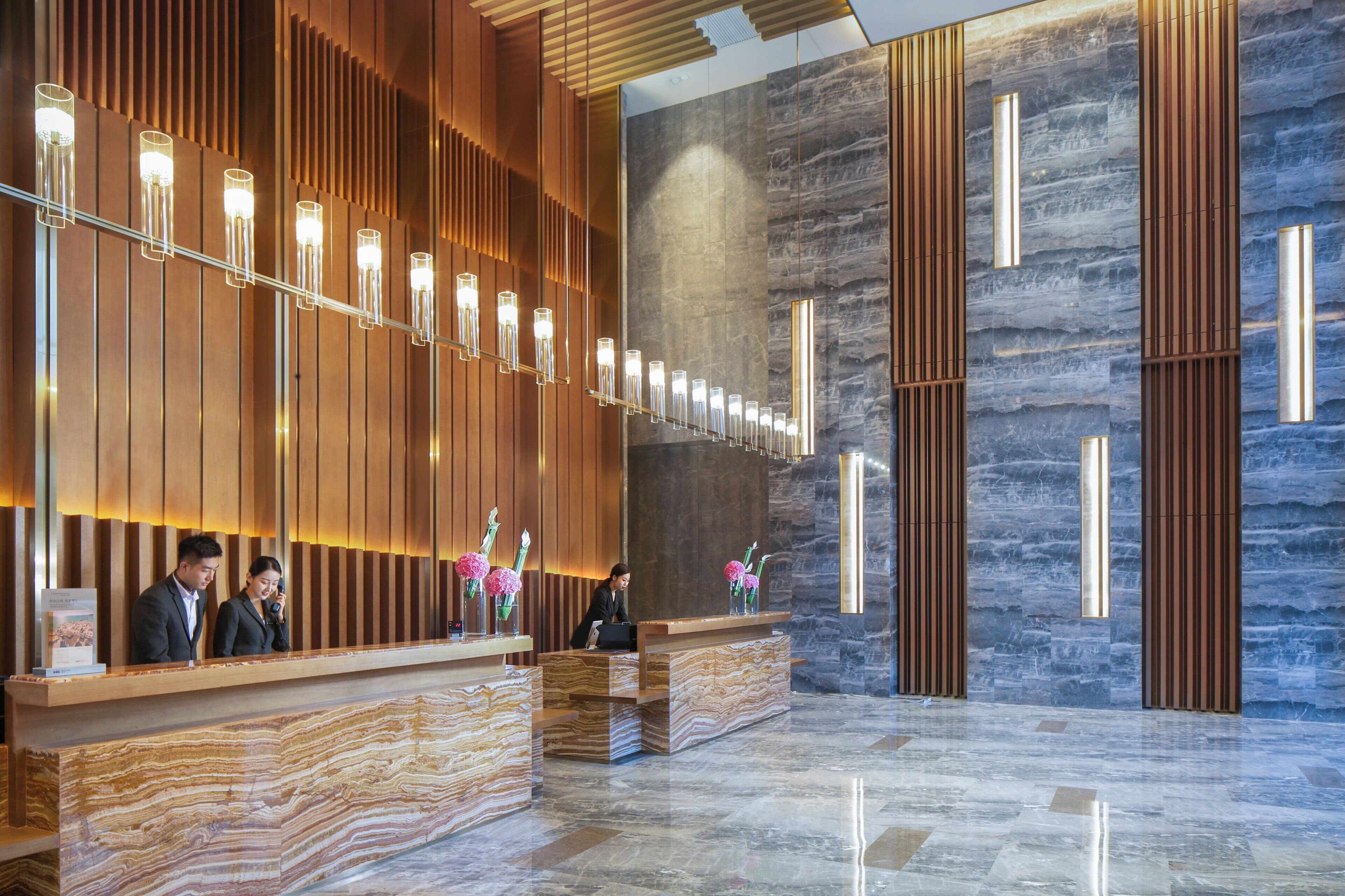 Zona de recepción de un hotel con un mostrador de madera, suelo de mármol y una pared moderna con listones verticales de madera y una sección de mármol.