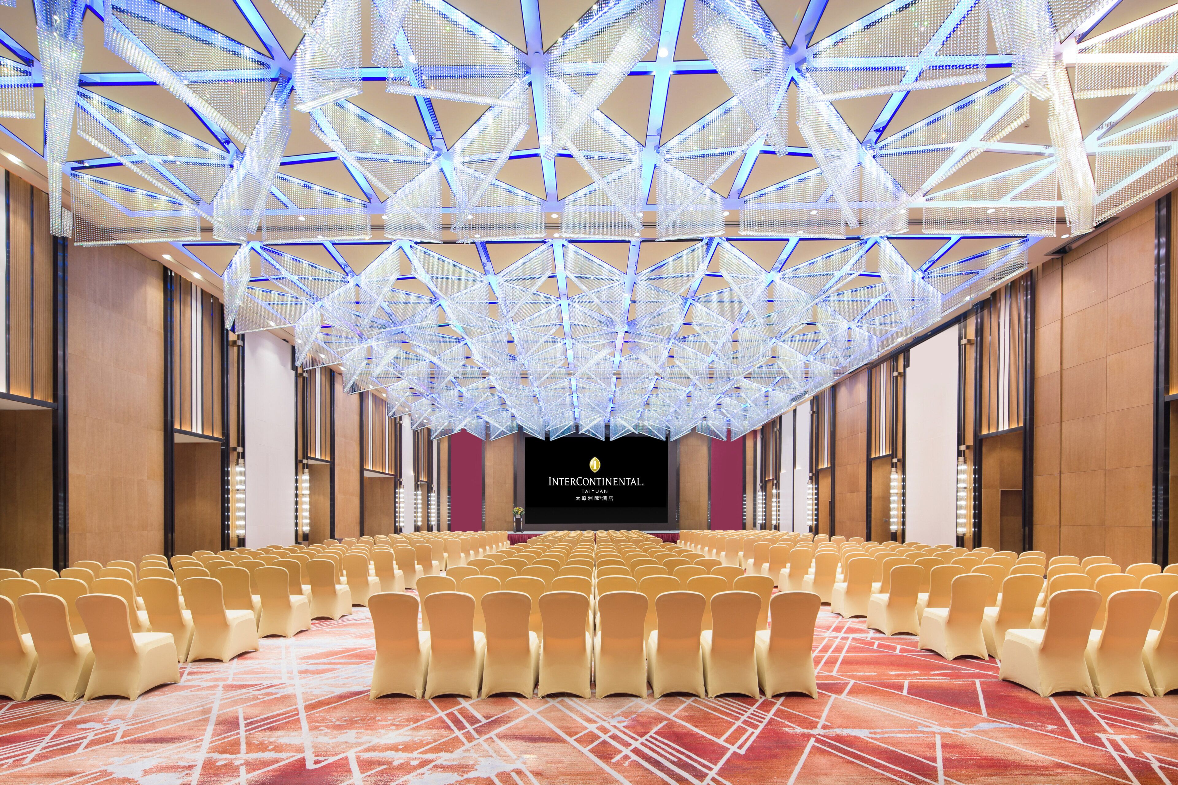A conference room with rows of chairs, a stage with a screen, and a ceiling with a geometric light fixture.