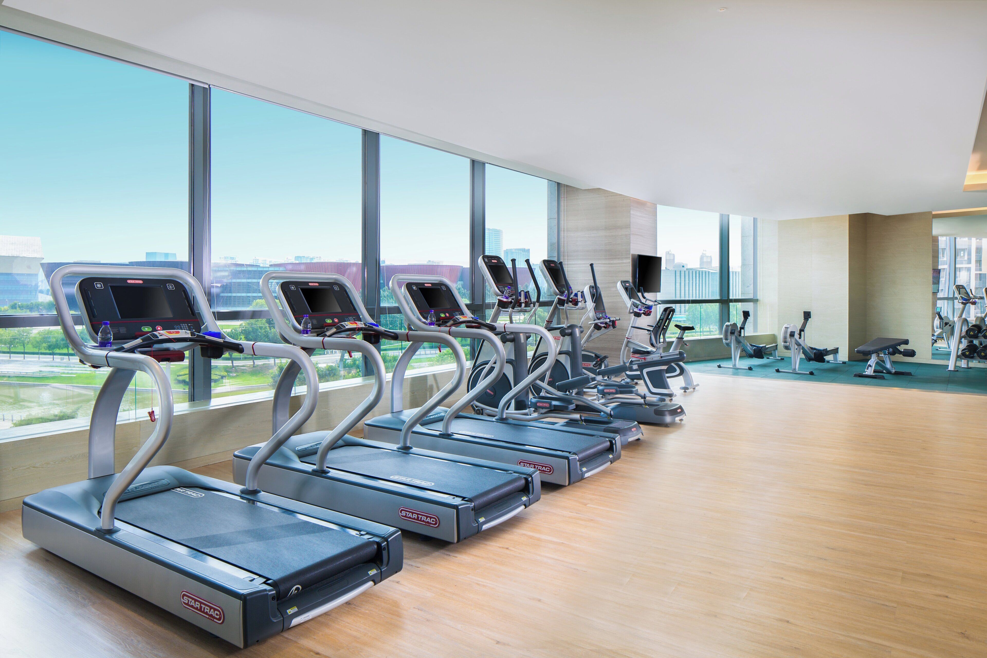 Ein Fitnessraum mit mehreren Laufbändern, die an einer Wand mit großen Fenstern aufgestellt sind.