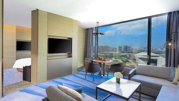 Une chambre d’hôtel moderne dotée d’une grande fenêtre donnant sur la ville, d’un téléviseur à écran plat fixé au mur, d’un canapé, d’une table basse et d’un coin repas avec des chaises.
