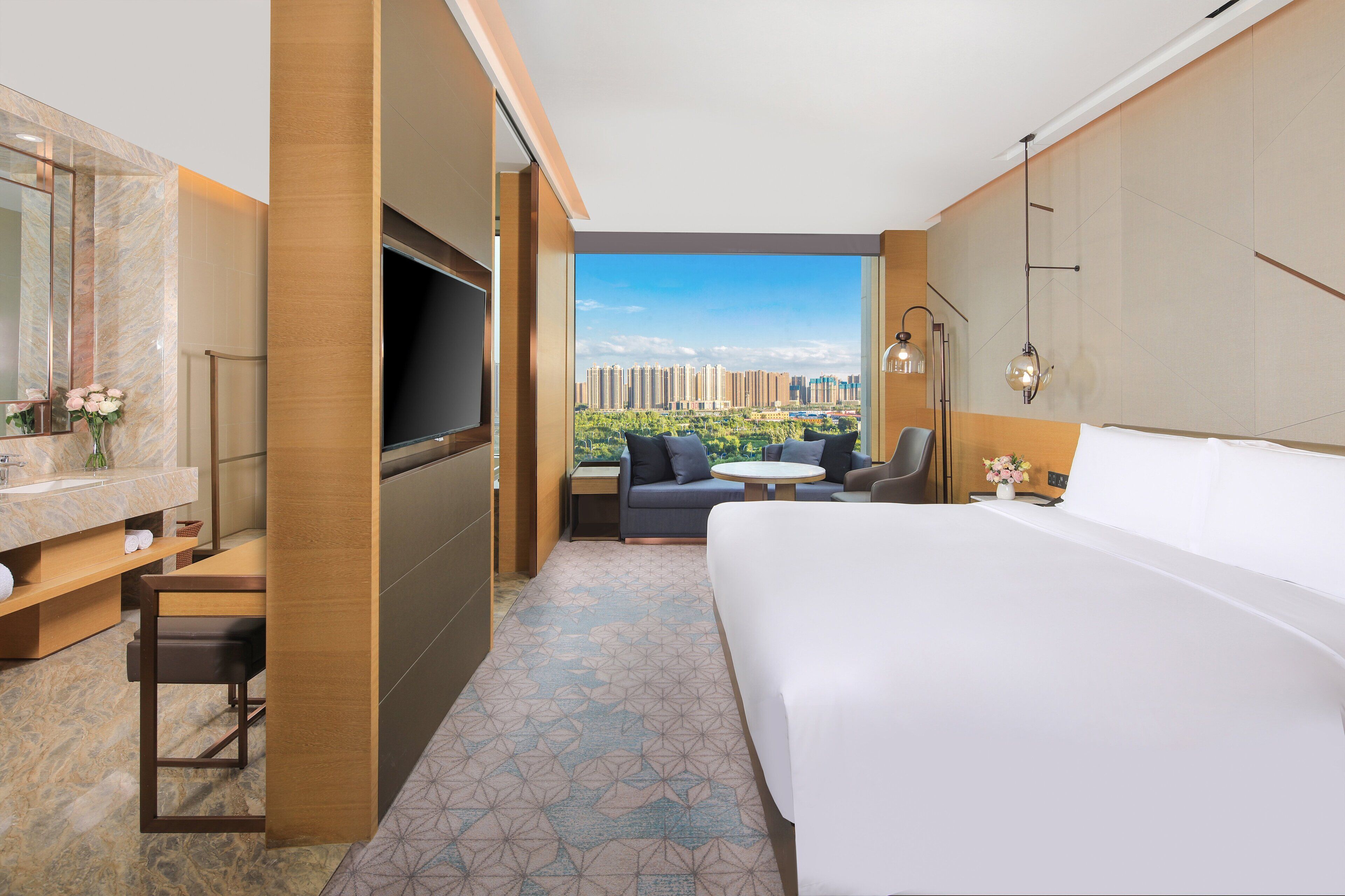 A hotel room with a large bed, a TV, a sofa, a small table, and a balcony with a city view.