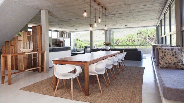Ruime eethoek met een lange houten tafel, witte stoelen, een houten bank en een bank met gekleurde kussens.