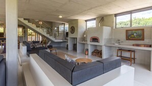 Een ruime woonkamer met een grote grijze hoekbank, een houten salontafel en een open haard. Een trap leidt naar een hoger gelegen verdieping.