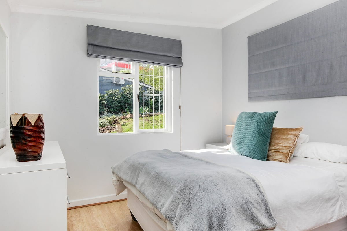 A bedroom with a bed, a bedside table, a window with a view of greenery, and a large grey headboard.