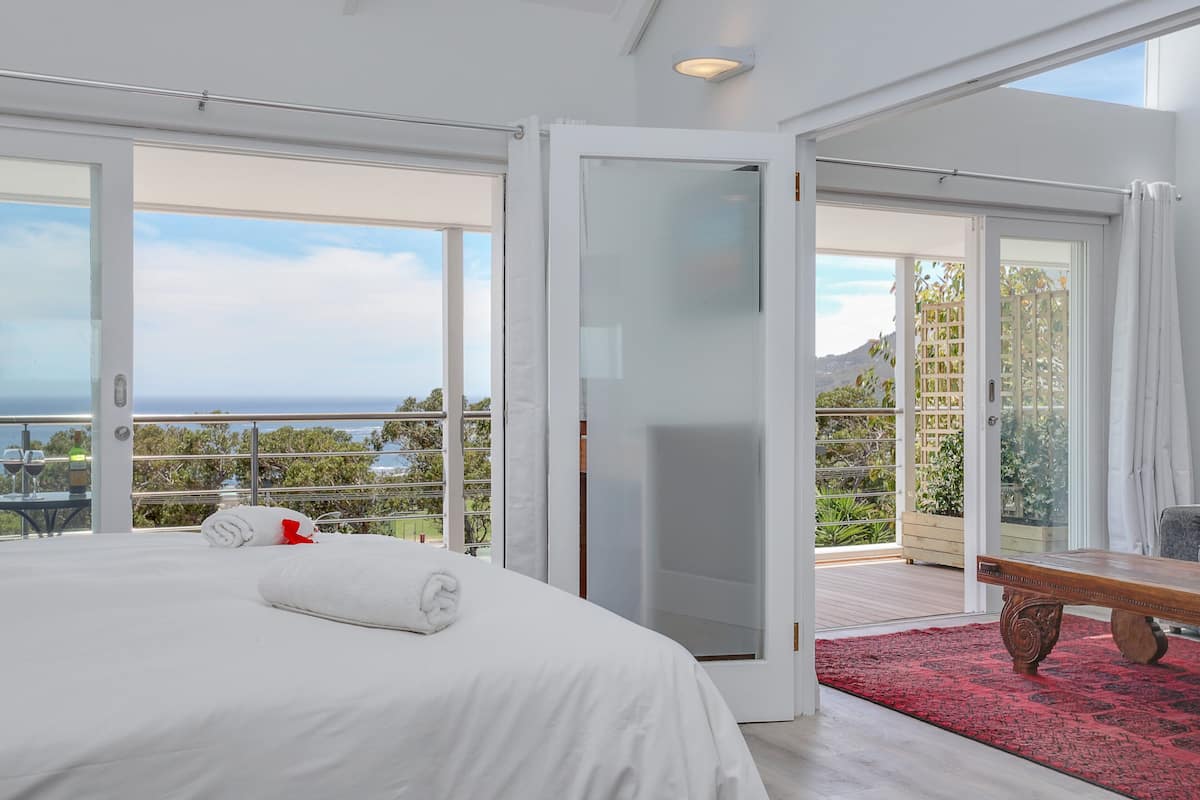 A spacious bedroom with a large bed, a wooden bench, and a red rug. It features a balcony with a view of the ocean and greenery.