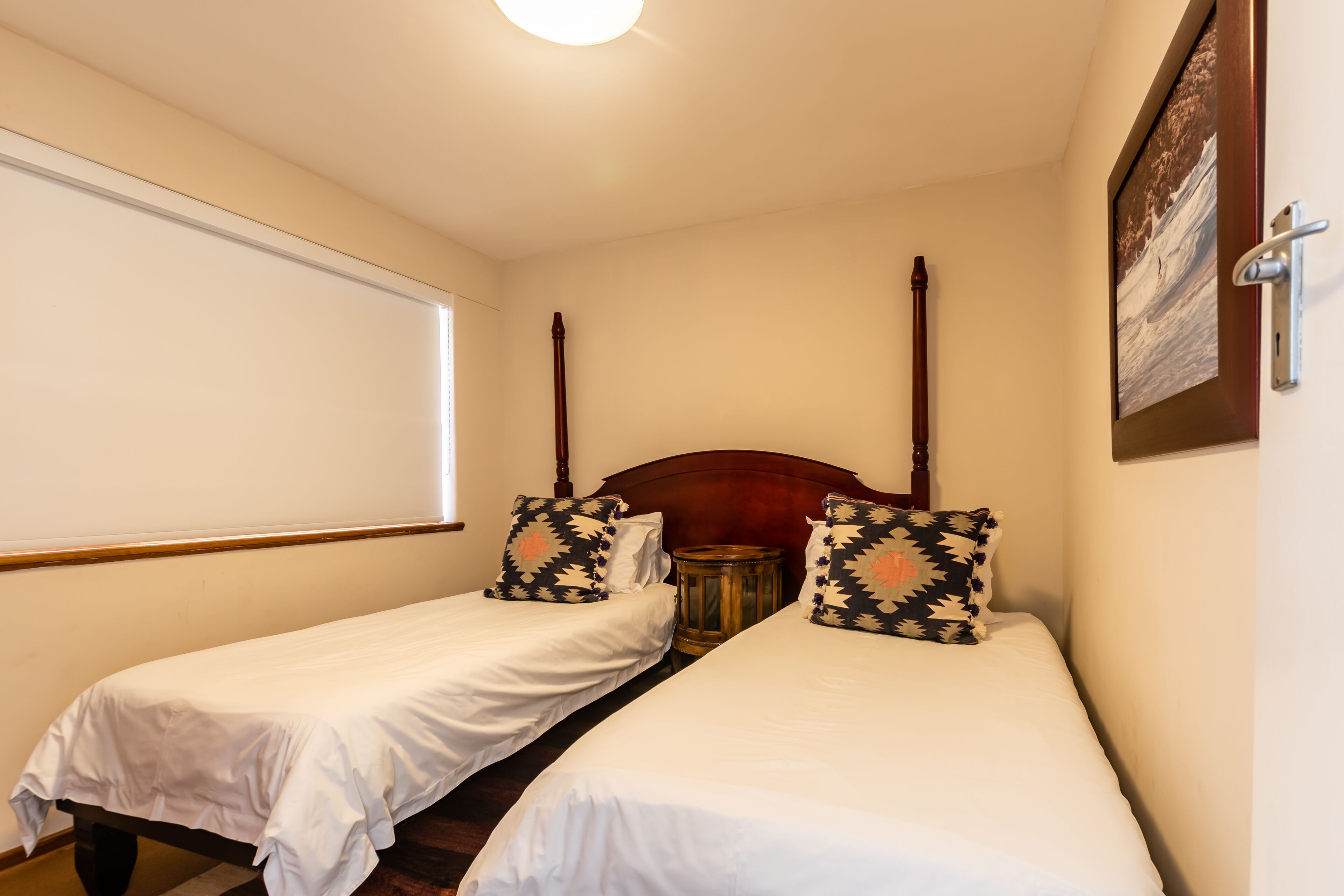 Una habitación con dos camas individuales, un cabecero de madera, una mesita redonda y una ventana con persianas.