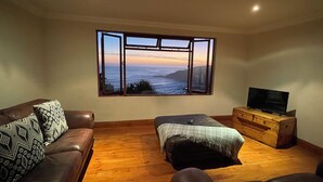 A living room with a large window, a sofa, an ottoman, a TV on a wooden dresser, and a wooden floor.