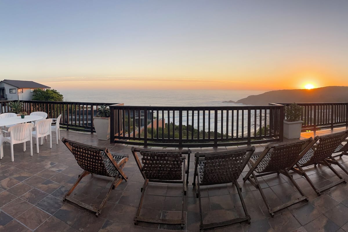 Une terrasse offrant une vue sur le coucher de soleil sur l’océan, aménagée avec des chaises et une table.