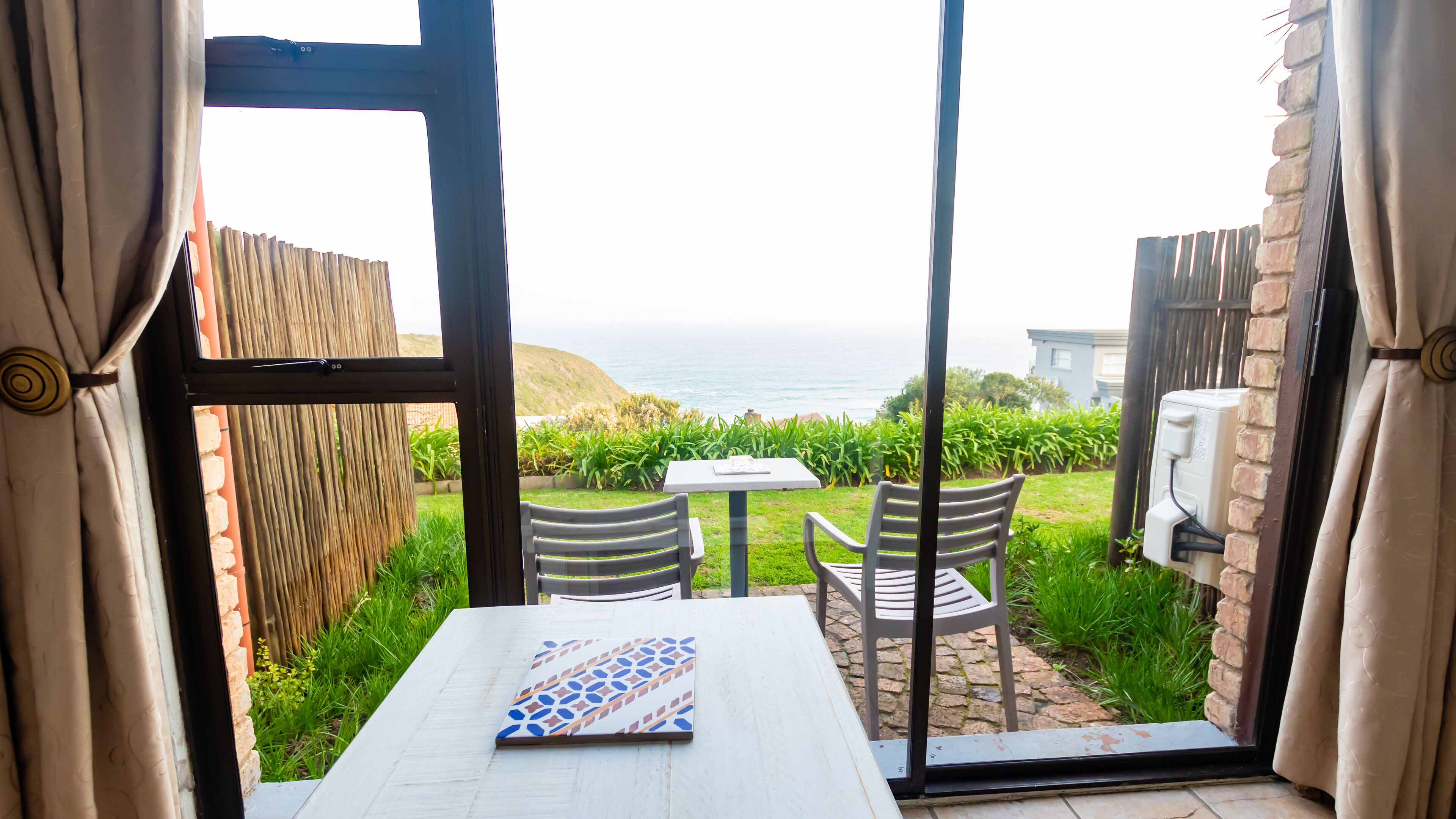 A room with a table and chairs, a view of the sea, and a garden outside.