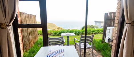 A room with a table and chairs, a view of the sea, and a garden outside.