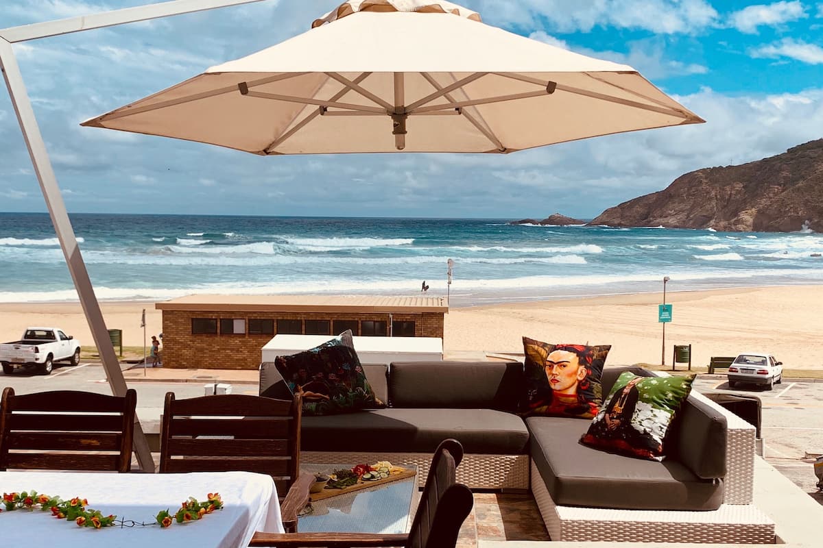 Un espace aménagé avec des tables, des chaises et un parasol, situé en bord de plage et offrant une vue sur l’océan.