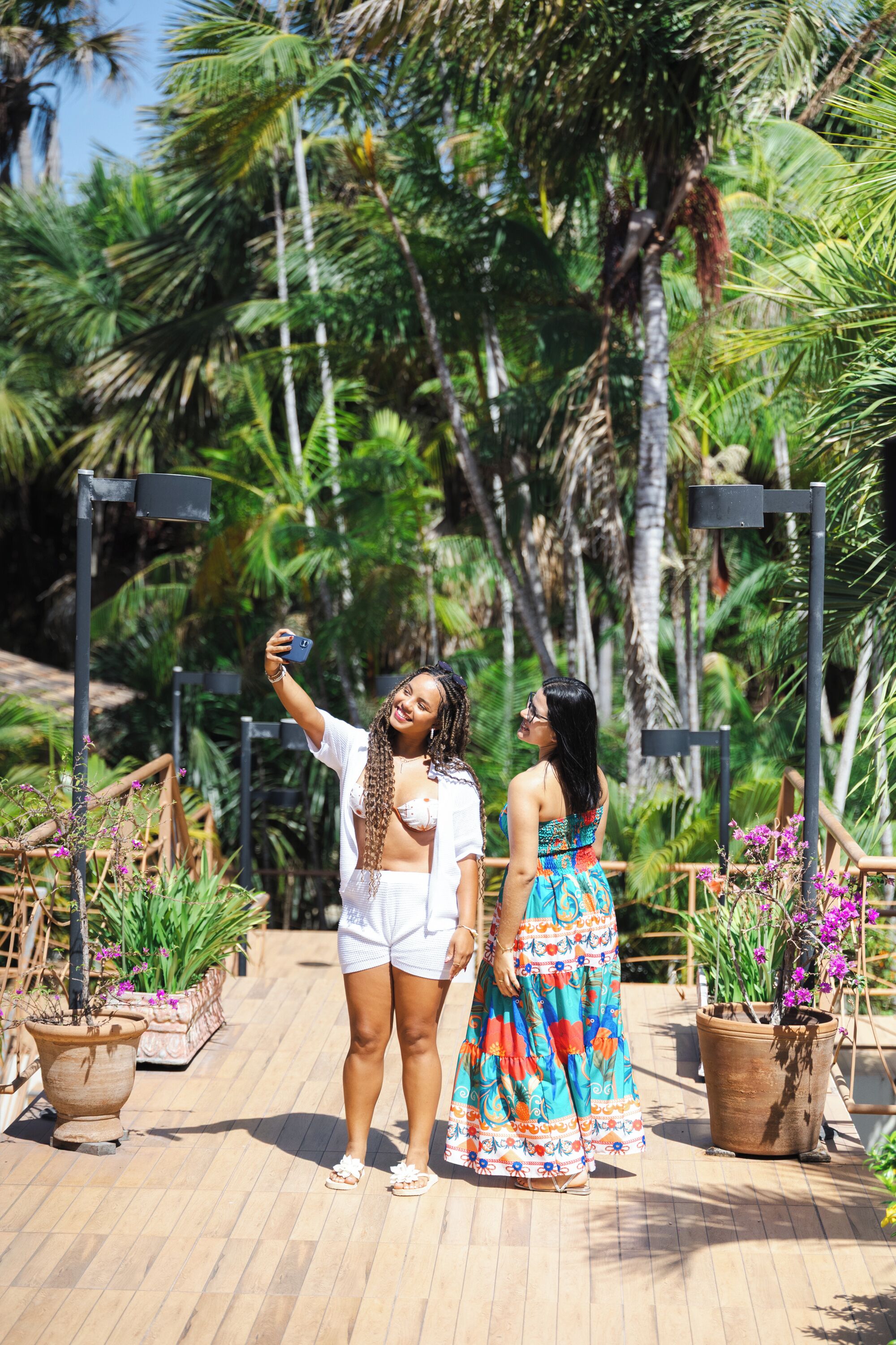 Two people taking a selfie in an outdoor setting with potted plants and palm trees.
