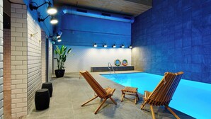 Zona de piscina interior con sillones de madera, una planta en maceta y un muro con azulejos azules.