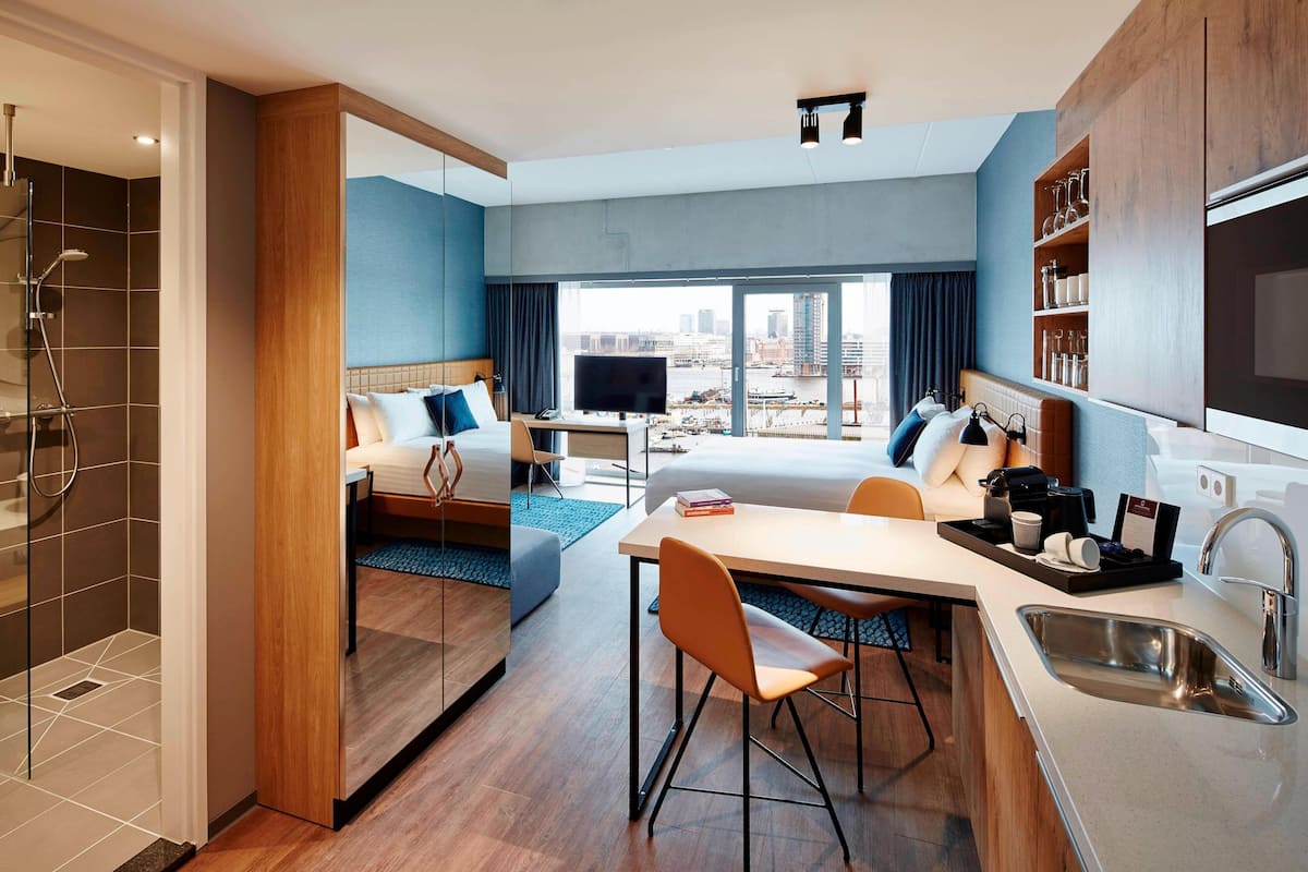 A modern hotel room with a bed, a desk, a dining area, a shower, and a view of the city.